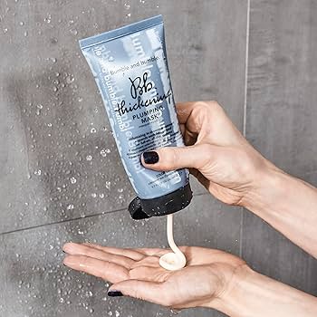 Hand holding a tube of Bumble and Bumble hair product with visible product being squeezed onto the palm. Bumble and Bumble Thickening Plumping Hair Mask 6.7oz jar, rich treatment that strengthens, nourishes, and adds body and volume to hair for fuller, thicker-looking strands.