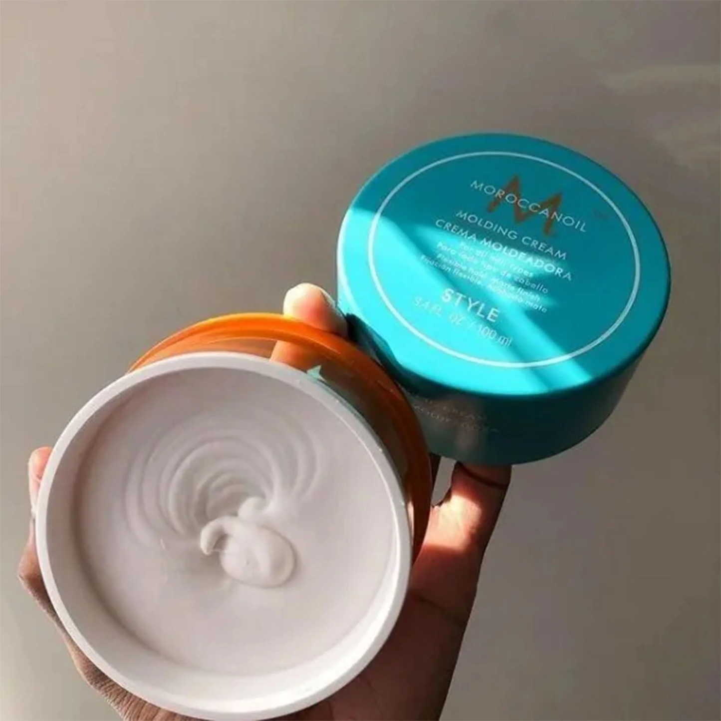 Hand holding a container of Moroccanoil styling cream with the lid open, showing the white cream inside.