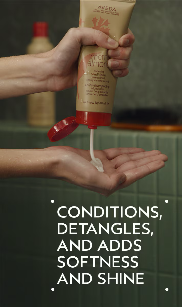 Person holding Aveda hair product with text about its benefits. Aveda Cherry Almond Softening Conditioner 6.7oz bottle, nourishing conditioner that softens hair, detangles, and leaves strands smooth, shiny, and healthy
