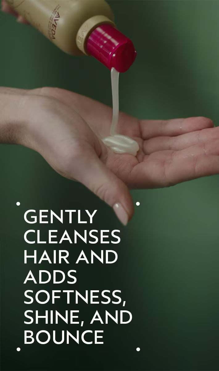 Hand holding a bottle of hair product with text on a dark background. Aveda Cherry Almond Softening Shampoo 8.5oz bottle, gentle shampoo that nourishes, softens, and leaves hair smooth, shiny, and healthy with a sweet cherry almond scent.