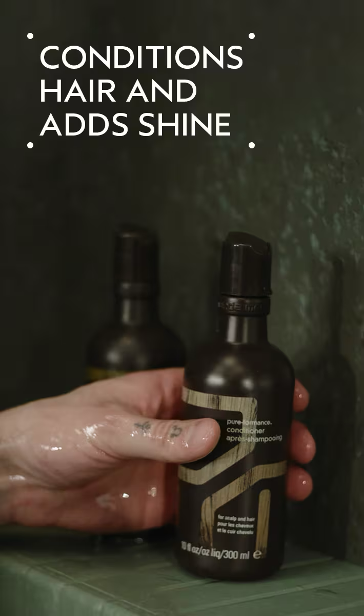 Hand holding a dark bottle with a label, against a dark background. Aveda Men Pure-Formance Conditioner 10oz bottle, moisturizing conditioner that soothes the scalp, reduces dryness, and leaves hair smooth and healthy.