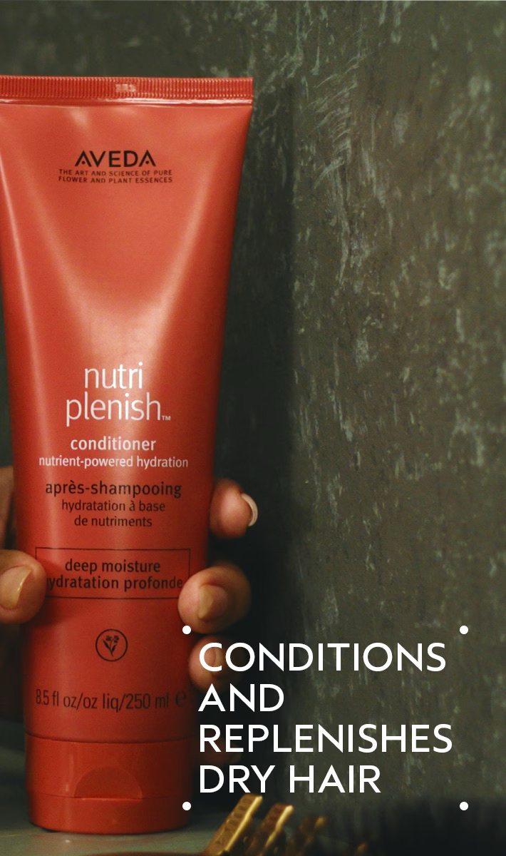 Red Aveda conditioner bottle held by a hand with text about hair care on a dark background. Aveda Nutriplenish Deep Moisture Conditioner 8.5oz bottle, rich conditioner that deeply hydrates, nourishes, and restores softness and shine to dry or damaged hair.