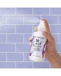 Hand holding a bottle of 'Bleach Free' conditioner against a tiled wall background. Bumble and Bumble Curl Reactivator 8.5oz bottle, lightweight spray that refreshes and reactivates curls, reduces frizz, and restores bounce, definition, and shine