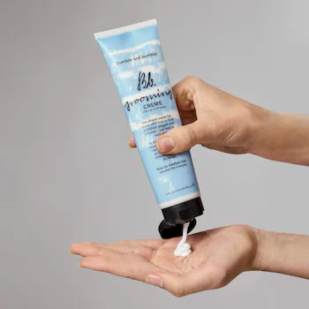 Hand holding a tube of Bumble and Bumble hair product with a small amount on the palm against a gray background. Bumble and Bumble Grooming Creme 5oz jar, versatile styling cream that provides light hold, smooths frizz, adds shine, and enhances manageability for all hair types