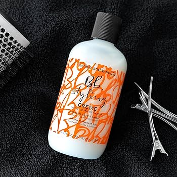 Bumble and Bumble styling crème bottle with a brush and clips on a black background. Bumble and Bumble Styling Creme 8.5oz jar, versatile styling cream that provides light hold, smooths frizz, adds shine, and enhances manageability for all hair types