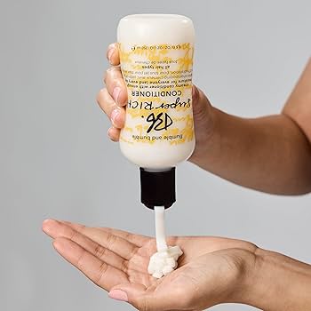 Hand holding a bottle of conditioner with a pump, dispensing some into another hand against a gray background. Bumble and Bumble Super Rich Conditioner 8.5oz bottle, deeply nourishing conditioner that hydrates, detangles, and softens hair while enhancing shine and manageability.