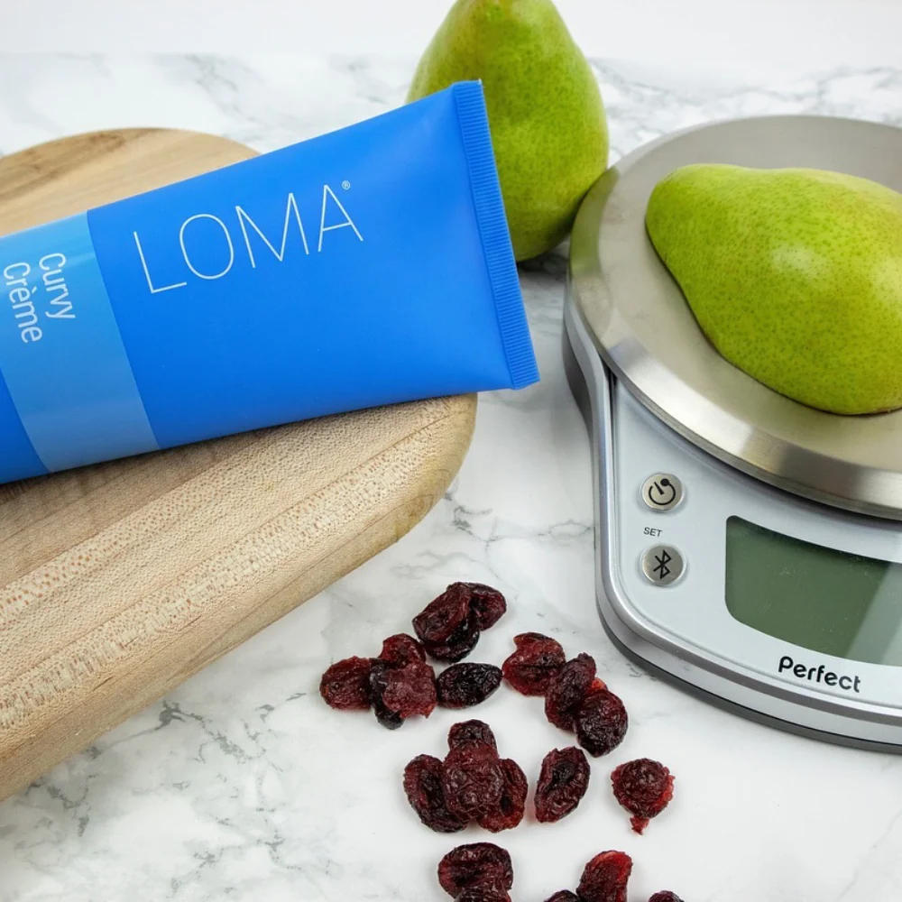 LOMA Creme packaging with a pear on a digital scale and dried berries on a marble surface. Loma Curvy Créme 8oz jar, styling cream that defines curls and waves, adds bounce and shine, controls frizz, and leaves hair soft and manageable.