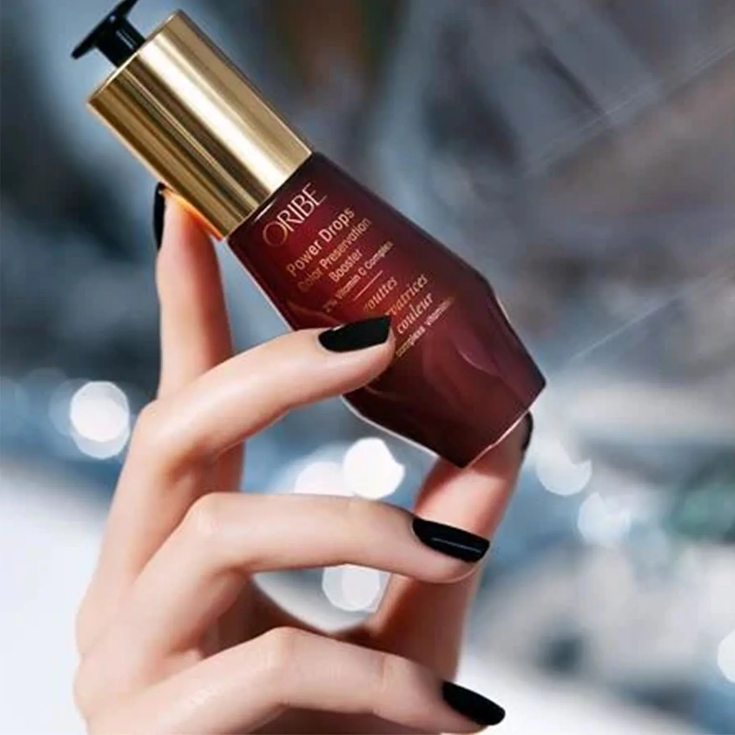 Hand holding a Oribe product with a blurred background. Oribe Color Power Drops Color Preservation Booster 1oz, 2% Vitamin C Complex that protects hair color, boosts vibrancy, and prolongs color-treated hair’s lifespan.