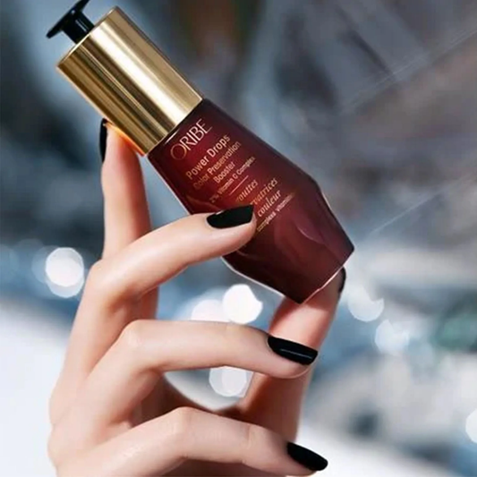 Hand holding a Oribe product with a blurred background. Oribe Color Power Drops Color Preservation Booster 1oz, 2% Vitamin C Complex that protects hair color, boosts vibrancy, and prolongs color-treated hair’s lifespan.
